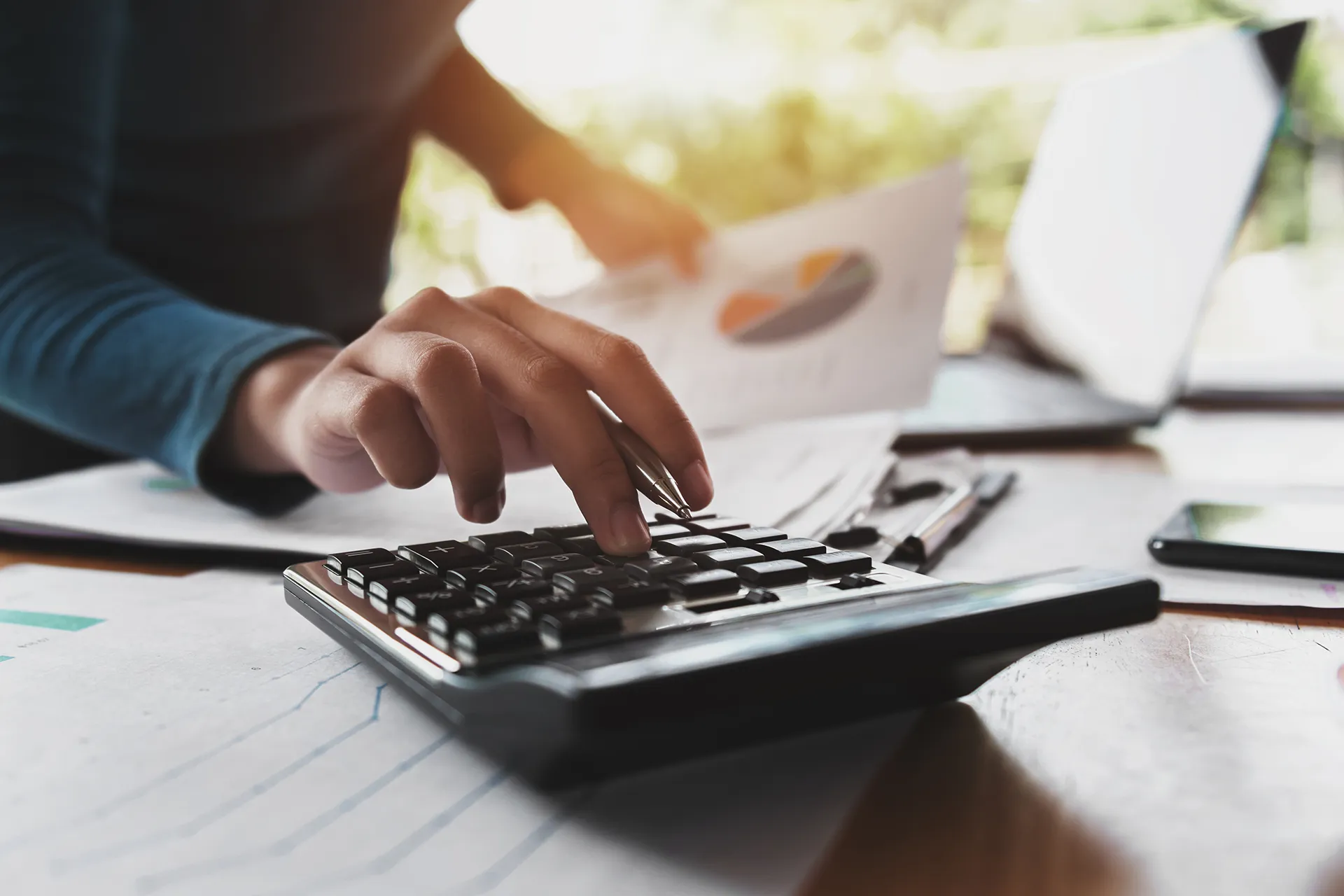 A person using a calculator to perform financial analysis while holding business documents featuring a colorful pie chart.