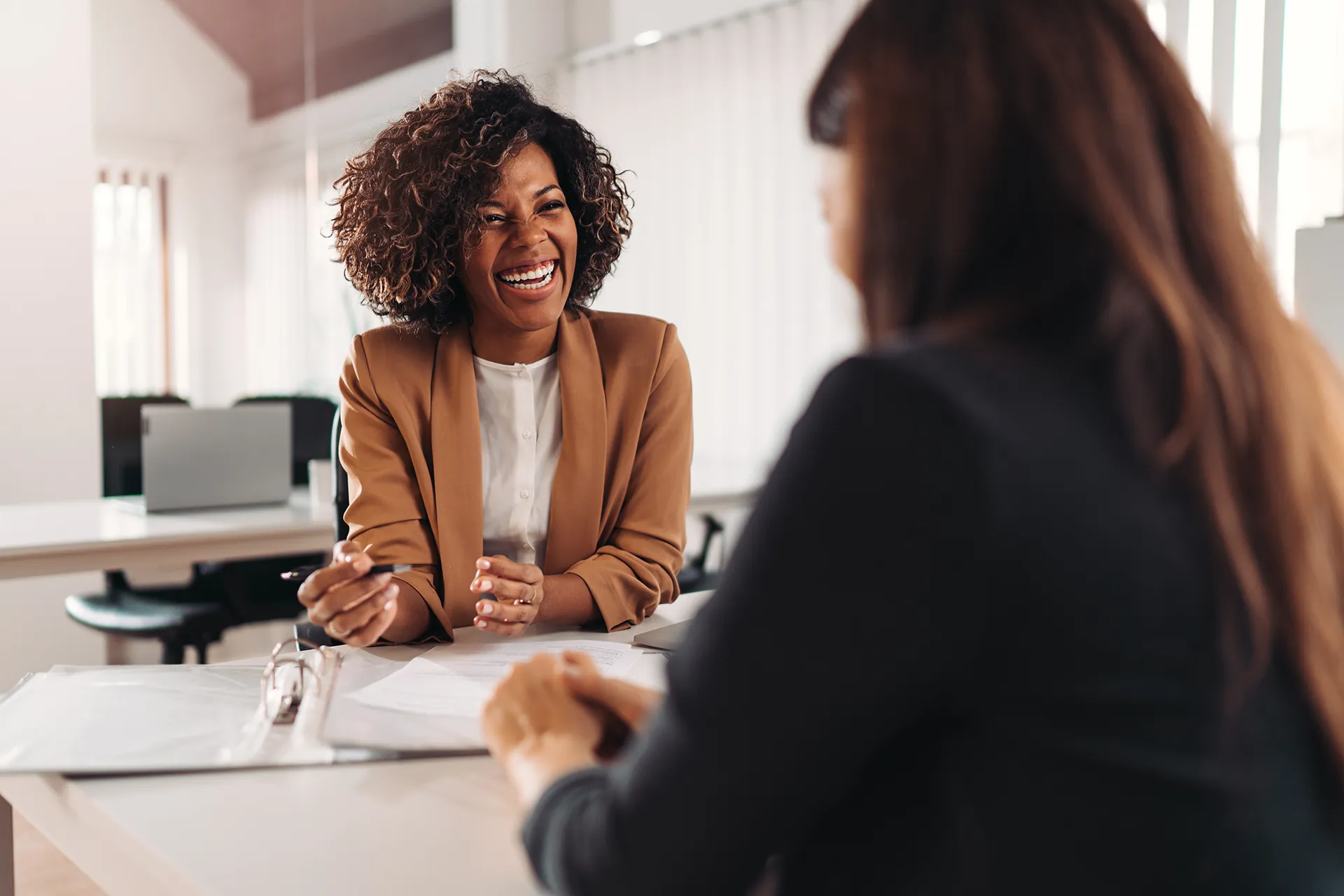 A smiling woman with curly hair sitting at a desk during a consultation, holding a pen and engaging in a positive discussion with a professional.