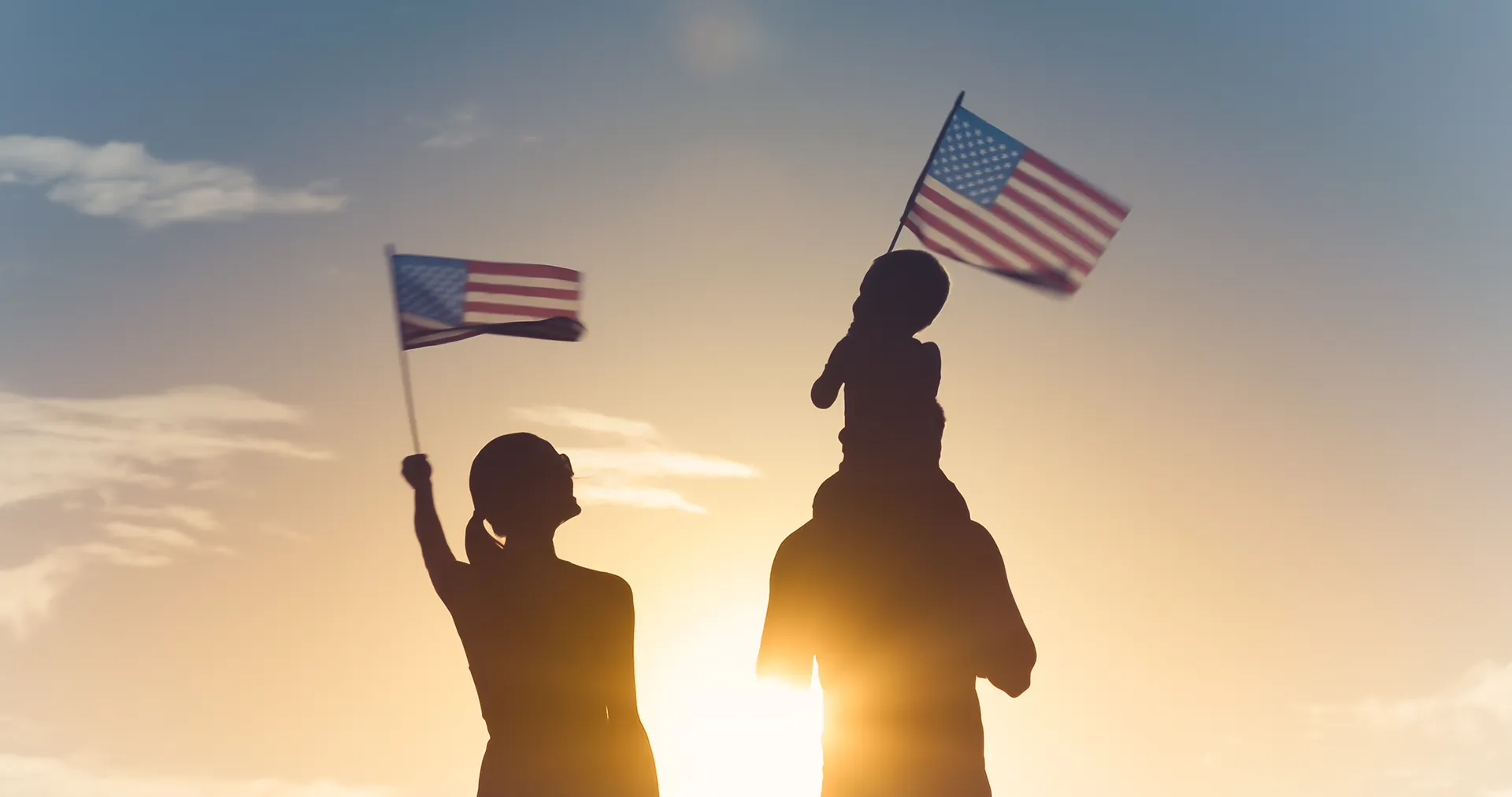 Silhouette of a family at sunset with a child on a parent's shoulders, all waving small American flags against a golden sky.